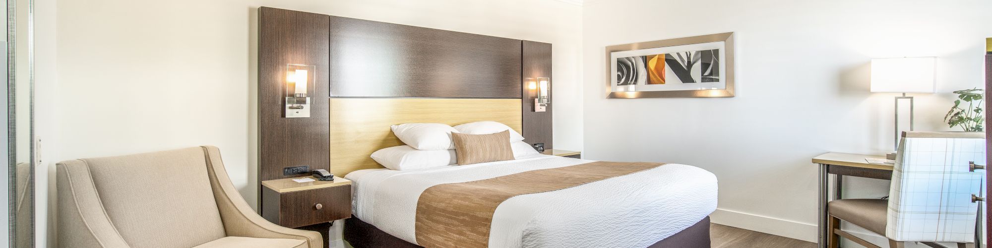 A neatly arranged hotel room with a large bed, an armchair, a desk, and a lamp. Light wooden flooring enhances the bright, inviting atmosphere.