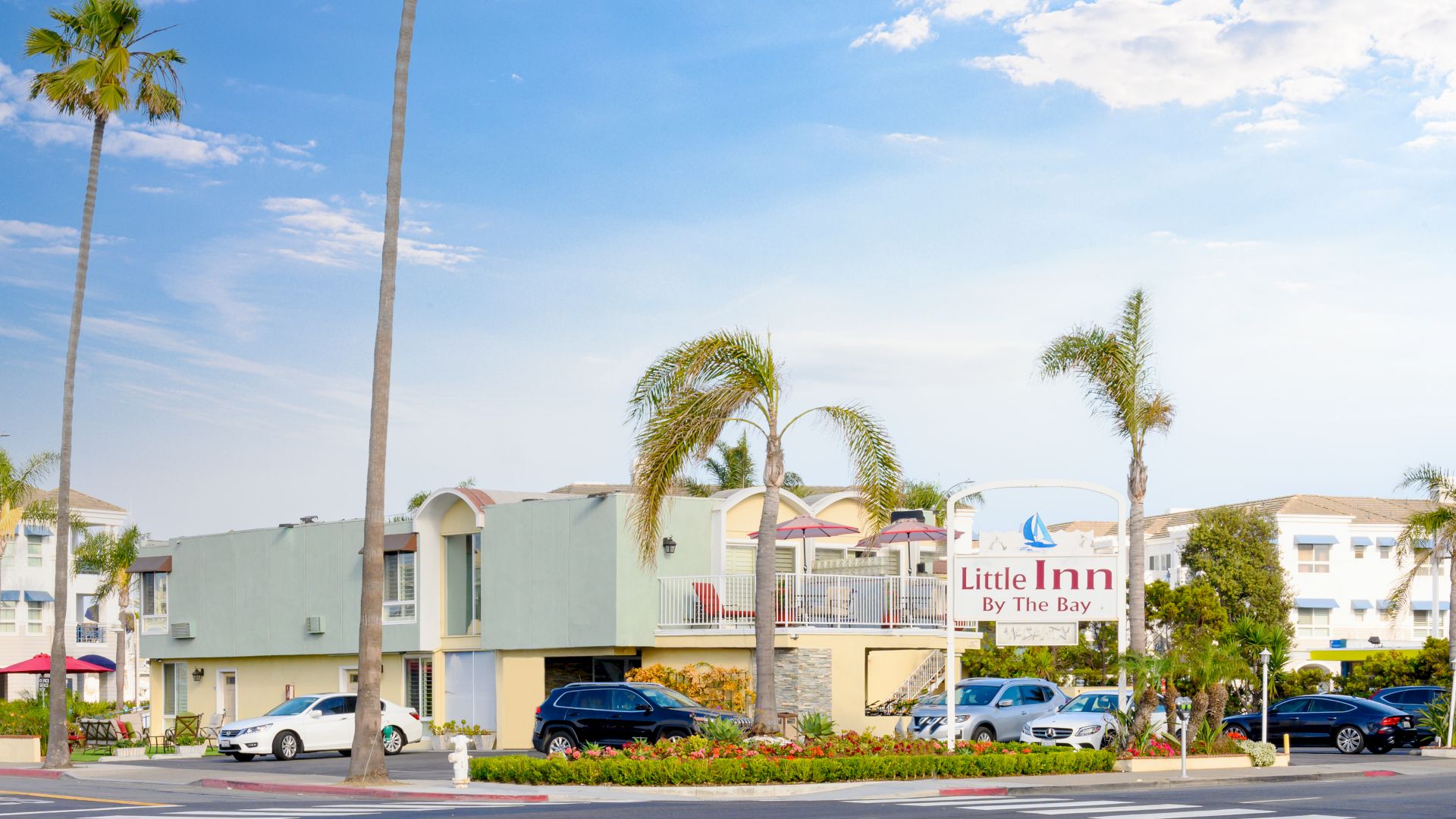The image showcases a hotel named "Little Inn By The Bay" with palm trees, parked cars, and a clear sky in the background.