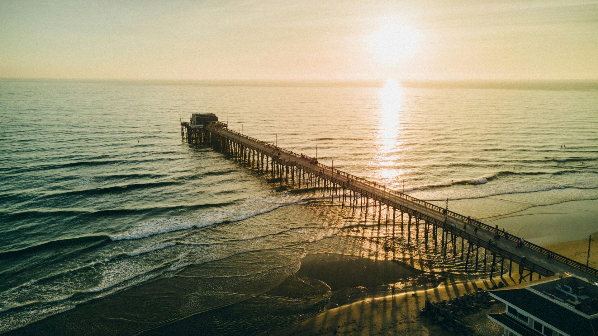 An aerial view of a long pier extending over the ocean with the sun setting on the horizon, casting a golden reflection on the water's surface.