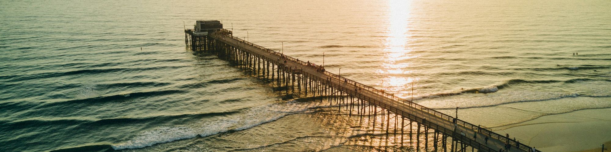 An aerial view of a long pier extending over the ocean with the sun setting on the horizon, casting a golden reflection on the water's surface.