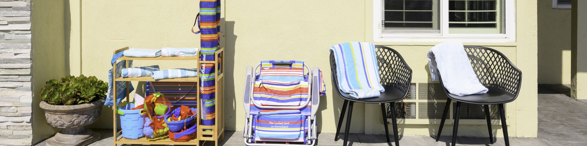 The image shows a patio with folded beach chairs, a rack of beach toys, and two black chairs with towels draped over them, in front of a yellow wall.