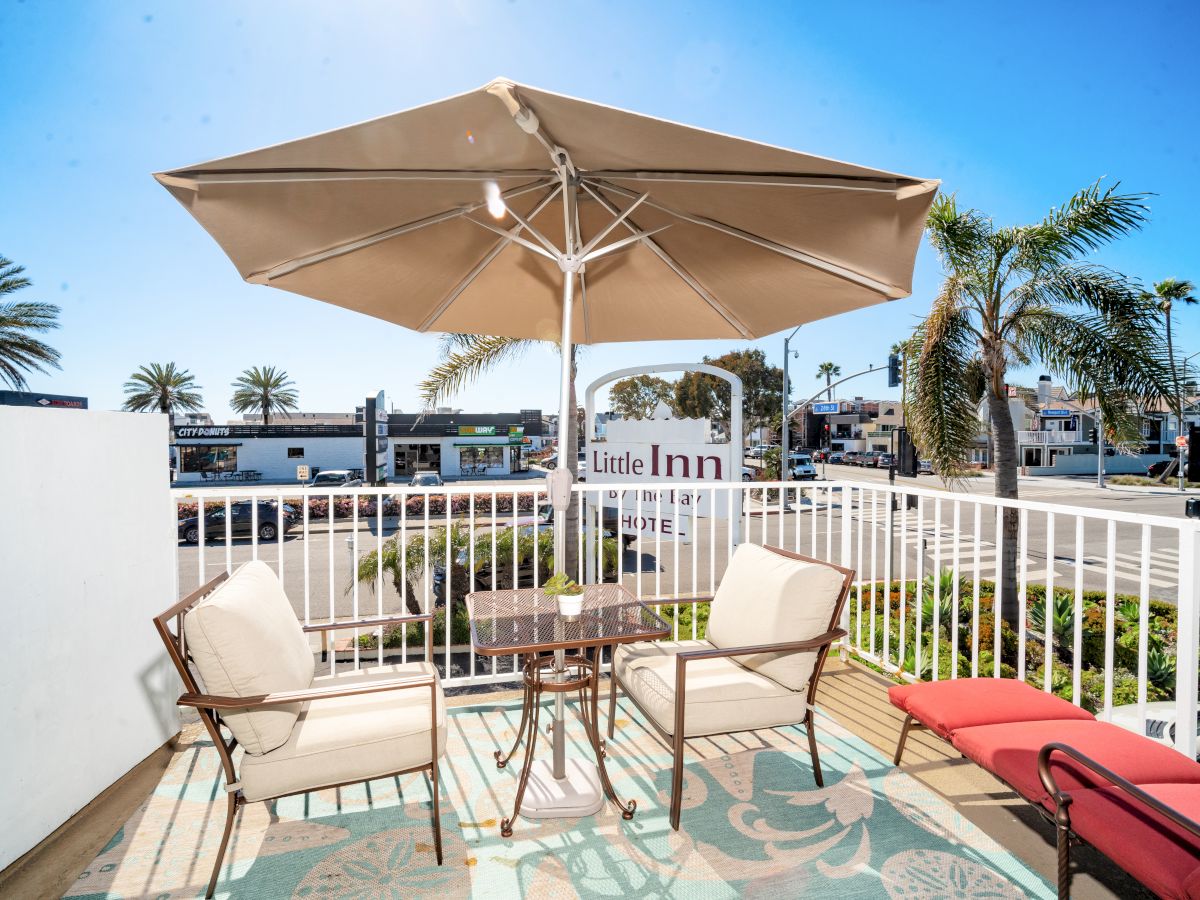 A patio with a table, chairs, and umbrella is shown, overlooking a street with palm trees and a sign reading 