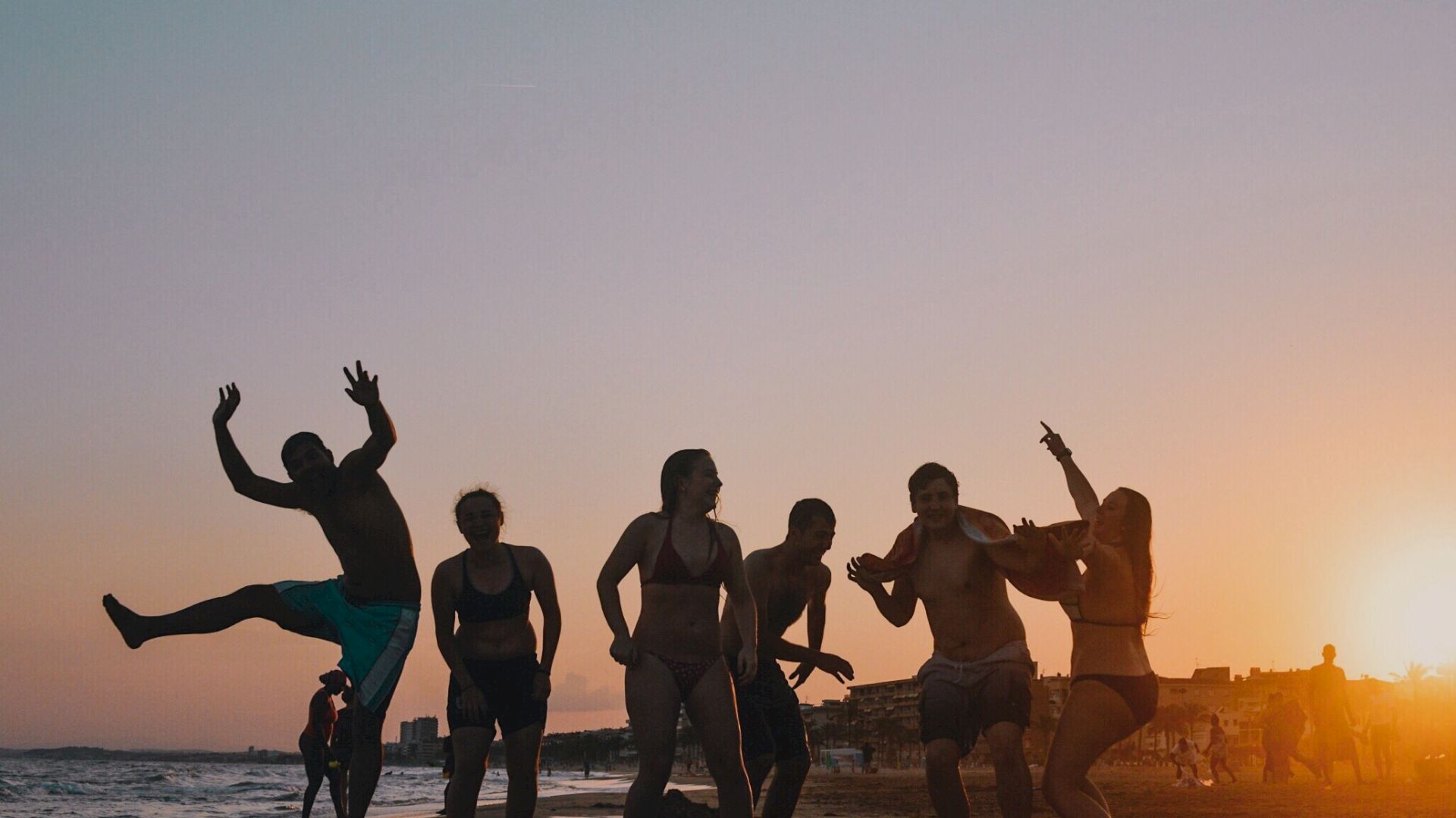 A group of people is having fun on the beach at sunset, with one person jumping joyfully.