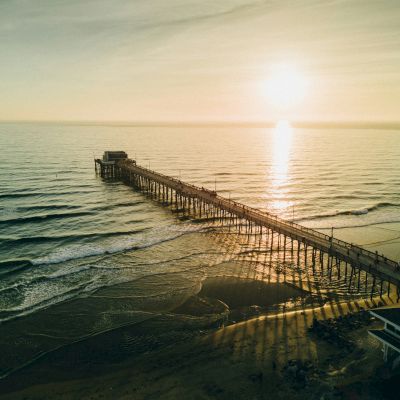 Aerial view of a long pier extending into a calm ocean during sunset, with waves gently rolling onto a sandy beach and a partially visible building.