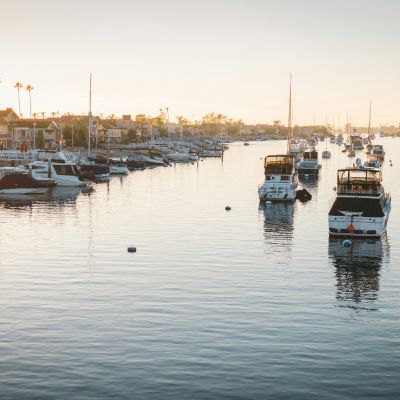 The image shows a serene marina at sunrise with several boats docked in the calm water, flanked by waterfront homes and palm trees reflecting in the water.