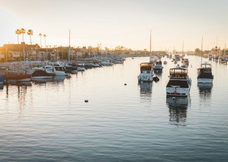 The image shows a serene marina at sunrise with several boats docked in the calm water, flanked by waterfront homes and palm trees reflecting in the water.