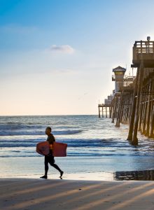 A surfer carrying a board walks along the beach as the sun sets behind a wooden pier extending into the ocean, casting long shadows on the sand.