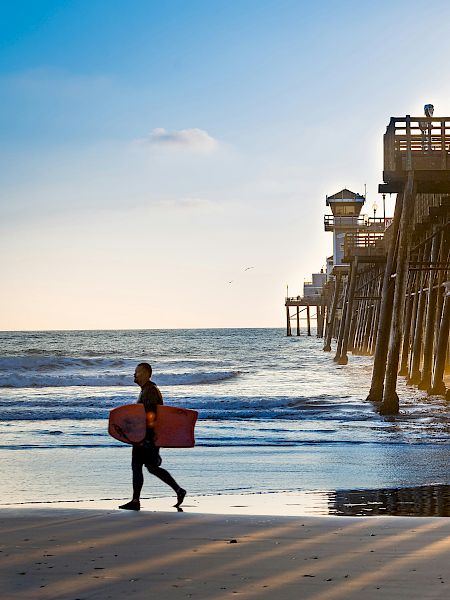 A surfer carrying a board walks along the beach as the sun sets behind a wooden pier extending into the ocean, casting long shadows on the sand.