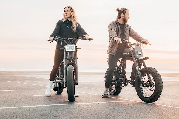 Two people are riding electric bikes by the seaside during sunset, looking in opposite directions and enjoying the scenery.