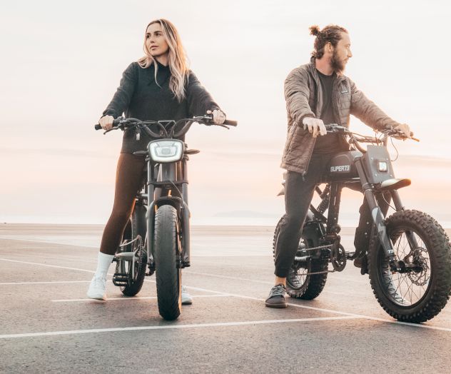 Two people are riding electric bikes by the seaside during sunset, looking in opposite directions and enjoying the scenery.