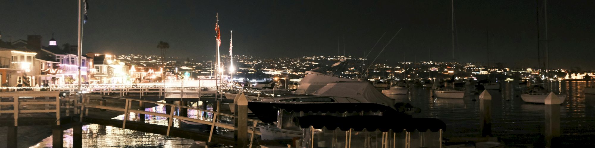 A nighttime harbor scene with boats docked, illuminated by city lights in the background. The scene reflects off the calm water.