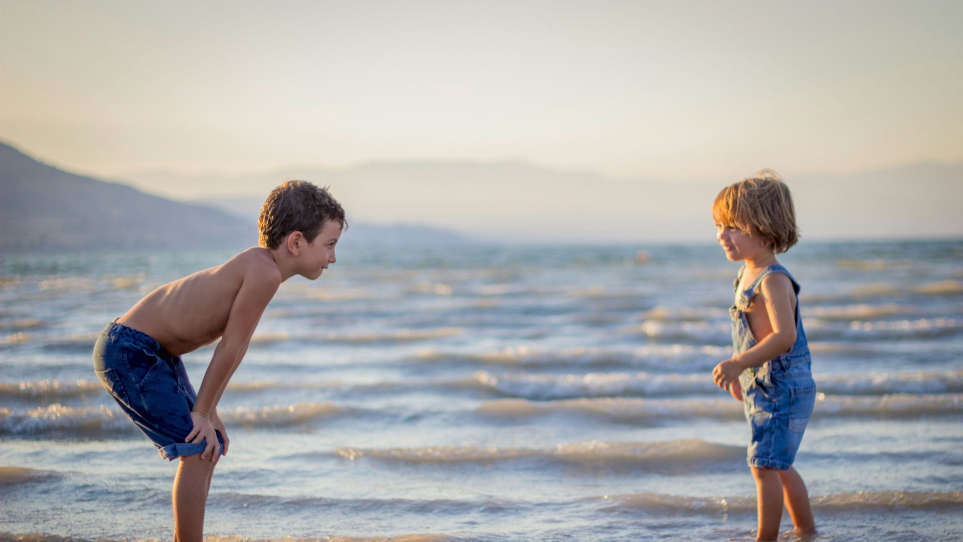 Two children are playing in shallow water at the beach during sunset, creating a playful and serene scene.