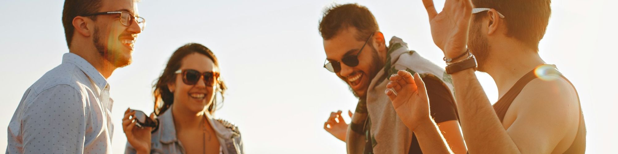 A group of four people stand outdoors in the sunlight, smiling and laughing together. They look like they are enjoying each other's company.