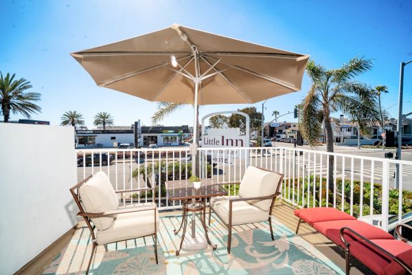 An outdoor patio with a table and chairs under an umbrella, adjacent to a street with palm trees and a sign that reads 