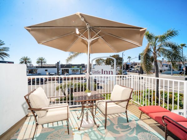 An outdoor patio with a table and chairs under an umbrella, adjacent to a street with palm trees and a sign that reads "Little Inn."