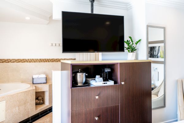 A modern hotel room features a mounted TV, a cabinet with a coffee station, a bathtub in the background, and a mirror on the wall.