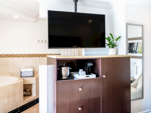 A modern hotel room features a mounted TV, a cabinet with a coffee station, a bathtub in the background, and a mirror on the wall.