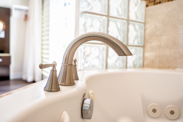 A close-up of a bathtub faucet in a bathroom, with glass block windows and a tiled wall in the background, and a blurred room on the left.