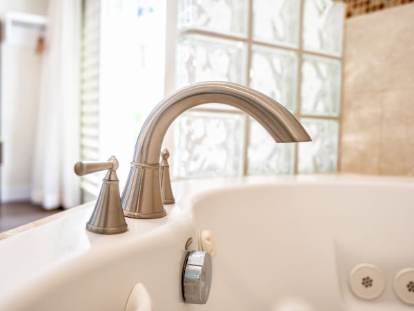 A close-up of a bathtub faucet in a bathroom, with glass block windows and a tiled wall in the background, and a blurred room on the left.