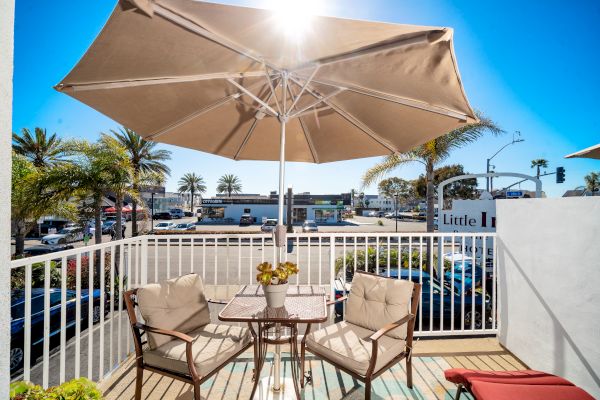 An outdoor patio with two cushioned chairs and a table under a large beige umbrella. The background reveals a sunny street with palm trees.