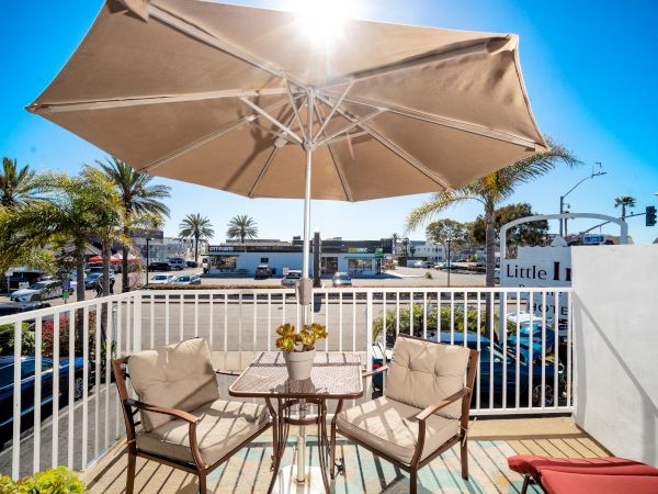 An outdoor patio with two cushioned chairs and a table under a large beige umbrella. The background reveals a sunny street with palm trees.