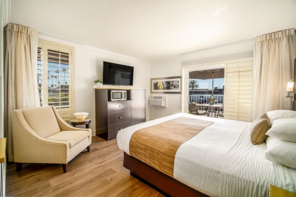 A cozy hotel room with a large bed, a TV atop a dresser, an armchair, and a balcony view. Light colors and natural light create a welcoming space.