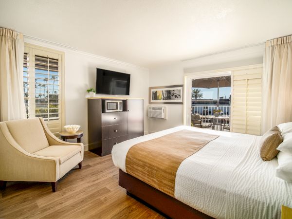 A cozy hotel room with a large bed, a TV atop a dresser, an armchair, and a balcony view. Light colors and natural light create a welcoming space.