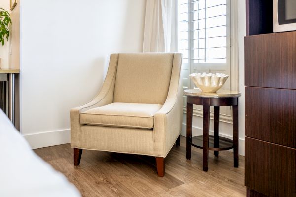 A beige upholstered armchair, a round side table with a decorative shell, and wooden flooring in a well-lit room.