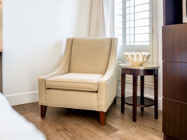 A beige upholstered armchair, a round side table with a decorative shell, and wooden flooring in a well-lit room.