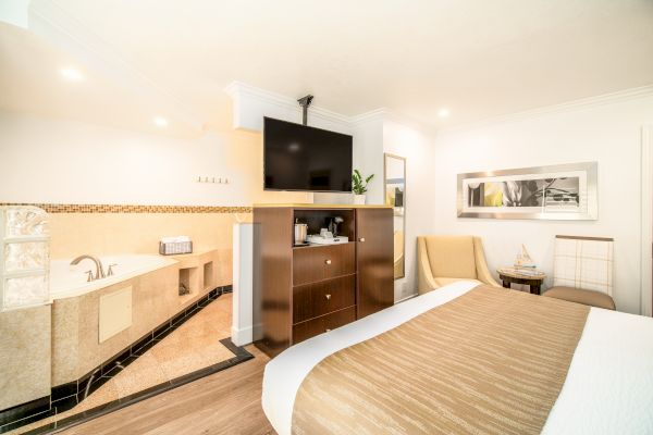 A modern hotel room features a bed, TV, jacuzzi, armchair, mirror, and a small table with seating.