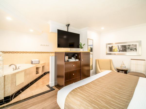 A modern hotel room features a bed, TV, jacuzzi, armchair, mirror, and a small table with seating.