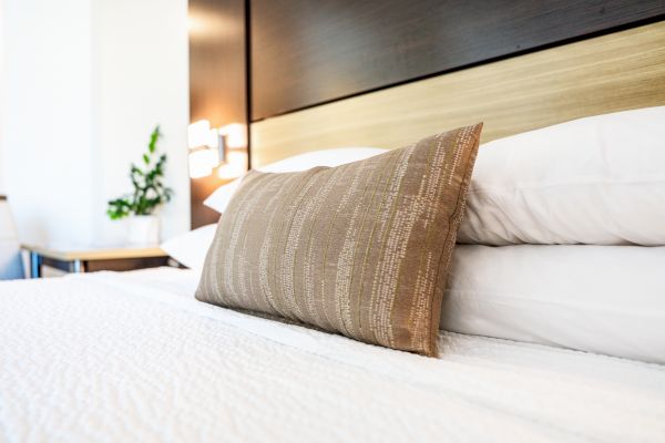 The image shows a neatly made bed with white linens and a decorative brown pillow. In the background, there is a wooden headboard and a small plant.