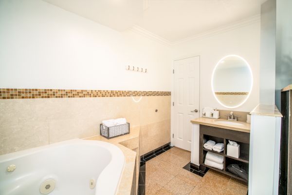 A bright bathroom with a large bathtub, a vanity with a round mirror, a sink, and a door. Towels are neatly arranged in a basket.