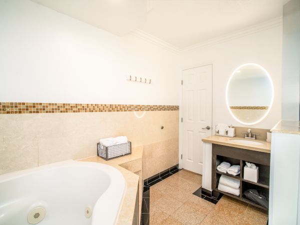 A bright bathroom with a large bathtub, a vanity with a round mirror, a sink, and a door. Towels are neatly arranged in a basket.