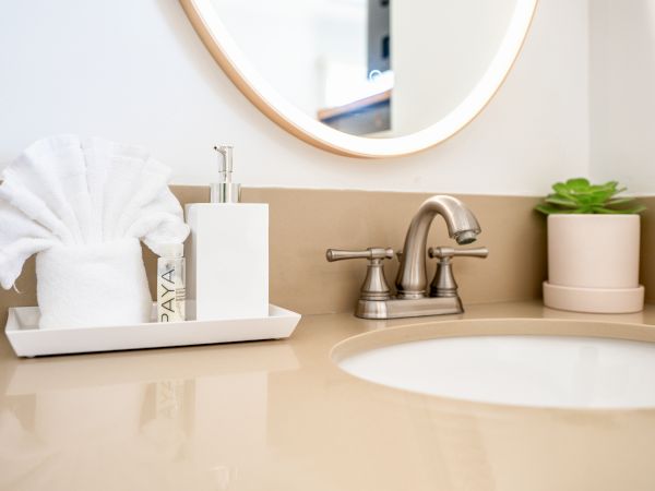 A bathroom sink with a beige countertop, a faucet, a mirror, a potted plant, a towel, a soap dispenser, and a bottle on a tray.
