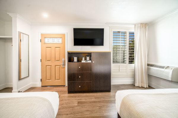 The image shows a hotel room with two beds, a wooden door, a TV mounted on the wall, a wooden cabinet, a window, and an air conditioner.