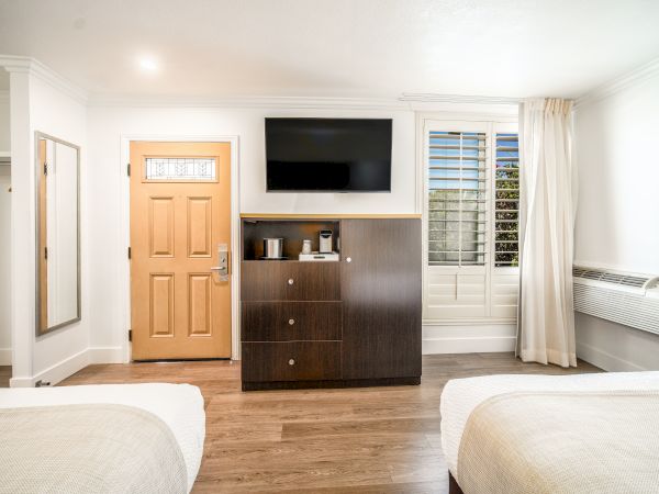 The image shows a hotel room with two beds, a wooden door, a TV mounted on the wall, a wooden cabinet, a window, and an air conditioner.
