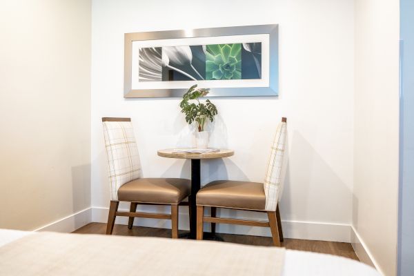 A cozy dining nook with two upholstered chairs and a small round table. A framed botanical art piece with green and white hues hangs above the table.