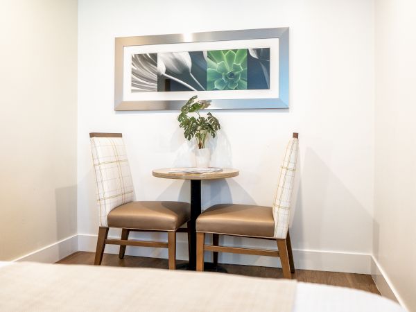 A cozy dining nook with two upholstered chairs and a small round table. A framed botanical art piece with green and white hues hangs above the table.