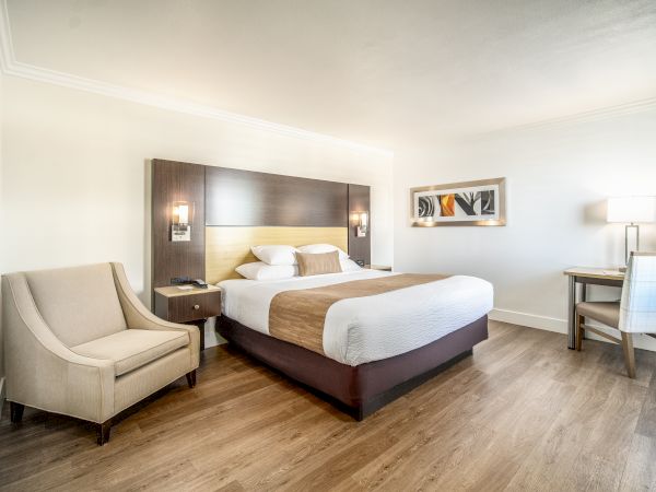 A modern hotel room with a king-size bed, wall art, bedside tables, an armchair, a desk with a chair, and a lamp on wooden flooring, ending the sentence.