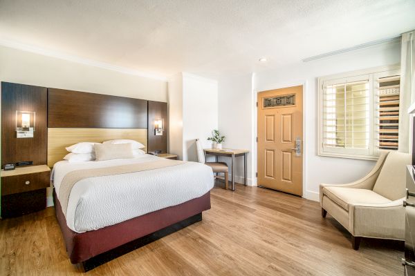 A modern hotel room with a large bed, desk, chair, and armchair, featuring wooden flooring and a well-lit, clean design.