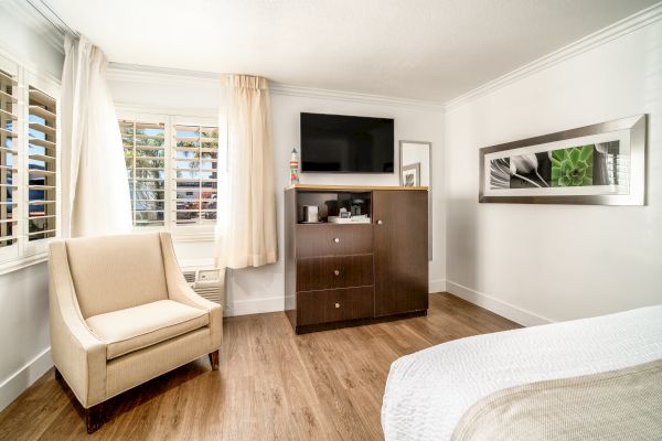 A cozy hotel room with a beige armchair, wooden flooring, a TV on a dresser, and wall art.