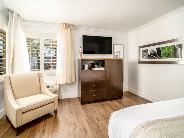 A cozy hotel room with a beige armchair, wooden flooring, a TV on a dresser, and wall art.