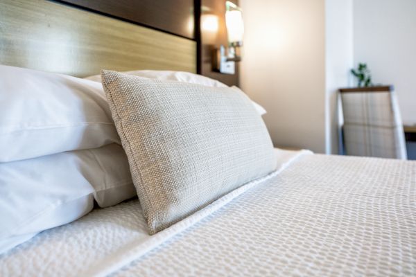 A neatly made bed with white linens, a textured beige pillow, a nightstand with a wall-mounted light, and a chair in the background.