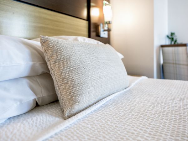 A neatly made bed with white linens, a textured beige pillow, a nightstand with a wall-mounted light, and a chair in the background.