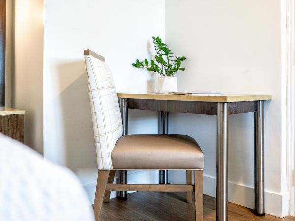 A small desk with a potted plant, a beige cushioned chair, and a corner of a room with white walls and a wooden floor.