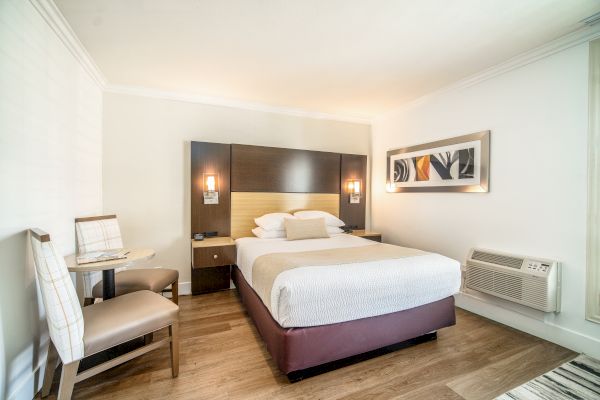 A neatly made bed in a modern hotel room, with wall artwork, two chairs and a small table, wooden flooring, and an air conditioner.