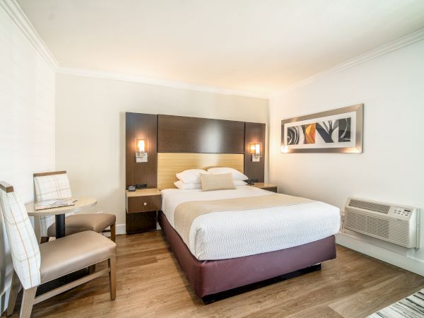 A neatly made bed in a modern hotel room, with wall artwork, two chairs and a small table, wooden flooring, and an air conditioner.