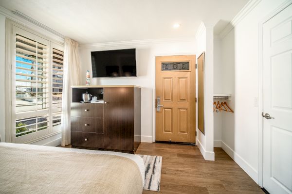 A modern hotel room features a bed, flat-screen TV, dresser, window with blinds, wall mirror, wooden door, and hangers on the right wall.