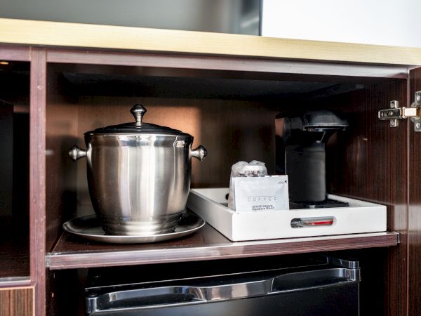 A cabinet with an open door reveals a metal ice bucket with a lid, a coffee maker on a tray with supplies, and a mini-fridge underneath.
